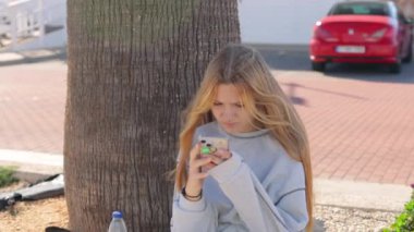 Elinde telefon olan genç bir kız caddede bir ağacın yanında oturuyor ve video kızın etrafında çekiliyor. Yüksek kaliteli FullHD görüntüler
