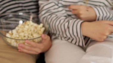 Çocuklar oturma odasındaki kanepede ellerinin üzerinde oturuyorlar. Erkek ve kız kardeş hafta sonları evde patlamış mısır yer ve birlikte televizyon izlerler. Erkek ve kız kardeş evde film izliyor.