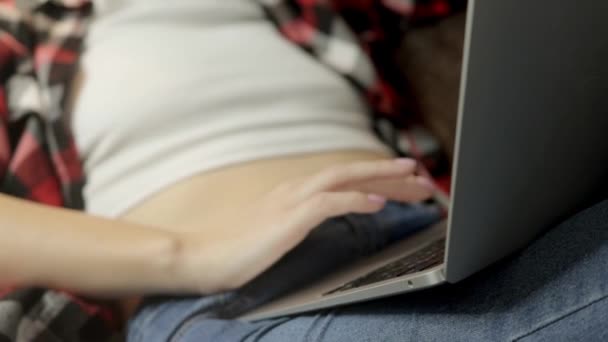 一个穿着红色衬衫和牛仔裤的女孩坐在客厅的沙发上 在她的电脑上工作 她全神贯注地浏览数字世界 展示了现代远程工作的概念 高质量的 — 图库视频影像