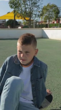 Sakallı küçük bir çocuk parkta oturup turuncu bir topla oynuyor.