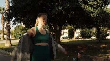 Spor giyimli genç bir kadın güneşli bir sabahta parkta egzersiz yapıyor. Kız esneme egzersizleri yapıyor..