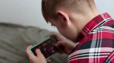 10 yaşında bir çocuk sabah kırmızı bir gömlek giyerek yatakta otururken akıllı telefondan video oyunu oynuyor. Yüksek kalite 4k görüntü