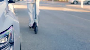Sokakta gri bir arabanın yanında scooterlı genç bir kızın görüntüsü. İnsanlar şehir caddesi boyunca elektrikli scooterlara biniyor. Yüksek kaliteli FullHD görüntüler