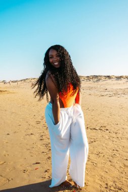 Kıvırcık gülümseyen Afro-Amerikan esmer tenli kadın beyaz pantolon ve mayoyla deniz suyunda yürüyor. Karışık ırk insanları, etnik köken, çeşitlilik.