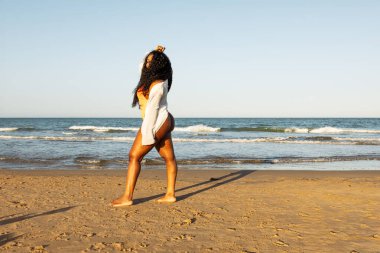Afro saç stili, kilolu ve obez bir kadının tam boy fotoğrafı. Plajda mayo ve beyaz gömlekle yürüyor. Koyu tenli kızın yaz tatili konsepti, vücut pozitifliği
