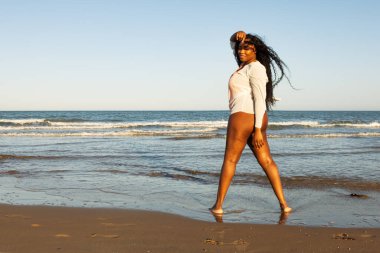 Turuncu bikinili güzel Afro-Amerikalı kadın ve tropik plajda beyaz gömlek. Denizde gülümseyen esmer kadın portresi. Mayolu esmer kız sahilde yürüyor ve eğleniyor..