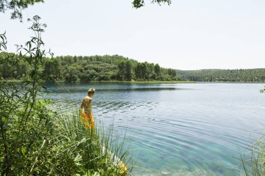 Bakir gölün kenarında duran genç adam, suyun sakinliği ve doğanın güzelliği tarafından büyülenmiş, bizi bekleyen ferahlatıcı yüzüşü ima ediyor.