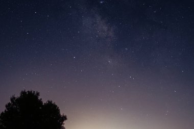 Samanyolu galaksisi yıldızlı bir gece gökyüzünde yayılıyor. Koyu maviden mor ve turuncuya bir eğim manzarayı tamamlıyor