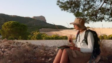 Şapkalı ve kitaplı bir kadın gezgin dağın kenarında oturur ve manzaranın tadını çıkarır.