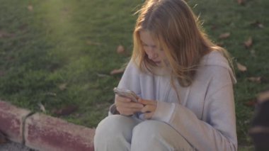 Elinde telefon olan genç bir kız parkta çimlerin üzerinde oturuyor. Sarışın, uzun saçlı, gündelik elbiseli bir kız. Yüksek kaliteli FullHD görüntüler