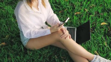 Genç bir kadın çimlerin üzerinde oturuyor ve dizüstü bilgisayarla çalışıyor..