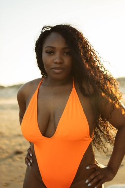 Kıvrımlı ve dalgalı bukleleri olan Afrikalı Amerikalı kadın canlı turuncu bir mayo giymiş sahilde poz veriyor.