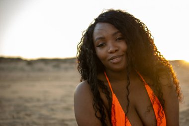 Kendinden emin, iri yarı bir kadın gün batımında kumsalda gülümsüyor. Havalı bir mayo giyip vücut pozitifliği yayıyor. Kaygısız bir yaz havasını somutlaştırıyor.