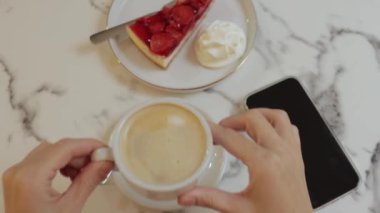 Akıllı telefon kullanan, elinde bir fincan kahve ve kek tutan bir kadın.