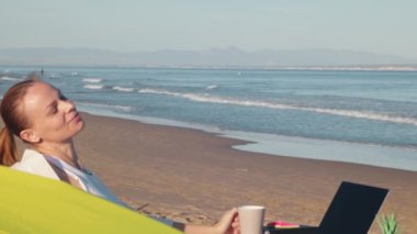 Genç bir kız sandalyede oturuyor ve dizüstü bilgisayar kullanıyor. Bir kız gün batımında sahilde çalışıyor..