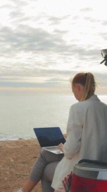 Dizüstü bilgisayarı olan genç bir kadın deniz kenarında oturmuş kitap okuyor..