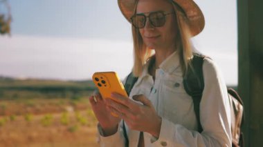 Şapkalı ve güneş gözlüklü genç bir kadın köy yolunda akıllı telefon kullanıyor.