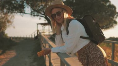 Sırt çantası ve hasır şapkalı genç bir kadın gün batımında parkta yürüyor.