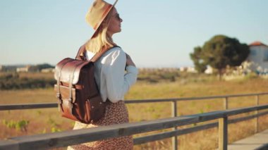 Sırt çantalı, hasır şapkalı genç bir kadın gün batımında köy yolunda yürüyor.