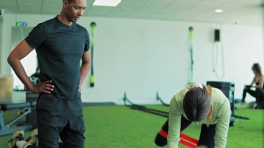 Spor salonunda antrenörle şınav çeken genç bir kadın.