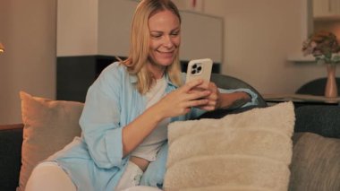 Oturma odasındaki koltukta cep telefonu kullanan genç sarışın kadın.