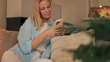 Evdeki rahat kanepede dinlenirken akıllı telefon kullanan mutlu genç bir kadın.