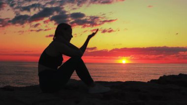 Siyah mayo giymiş bir kızın silueti gün batımında sahilde oturuyor.