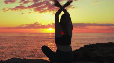Genç ve güzel bir kadın gün batımında deniz kenarında yoga yapıyor.