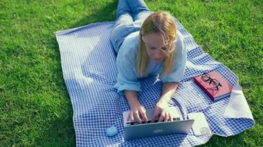 Ekose gömlekli genç bir kadın çimlere uzanıp defter okuyor.