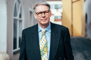 Koyu takım elbiseli ve gözlüklü şık bir iş adamı. Şehir manzarası da arkasındaymış.