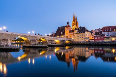 Regensburgs taş köprüsünün nefes kesen manzarası Tuna Nehri 'ni aydınlatırken Bavyera şehrinin gökyüzü akşam ışığında parlarken yansıtıyor.