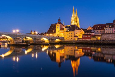 Regensburgs taş köprüsünün nefes kesen manzarası Tuna Nehri 'ni aydınlatırken Bavyera şehrinin gökyüzü akşam ışığında parlarken yansıtıyor.