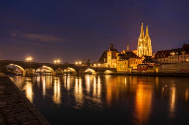 Regensburgs taş köprüsünün nefes kesen manzarası Tuna Nehri 'ni aydınlatırken Bavyera şehrinin gökyüzü akşam ışığında parlarken yansıtıyor.