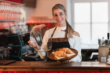 Sarışın bir garson bir kahve dükkanında bir tepsi kek tutuyor ve çörekleri müşteriye sunuyor.