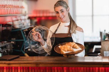 Sarışın bir garson bir kahve dükkanında bir tepsi kek tutuyor ve çörekleri müşteriye sunuyor.