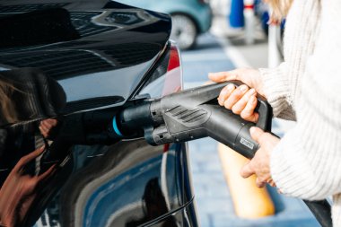 Bir kadın şarj aletini şarj istasyonundan siyah elektrikli arabasının prizine takıyor.