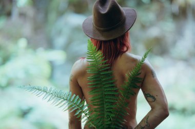 Sırtında yeşil eğrelti otu yaprakları ve kahverengi şapkalı kızıl saçlı kadın. Doğada ve arkadan fotoğraflanmış. Sağlık ve doğa projeleri kavramı.