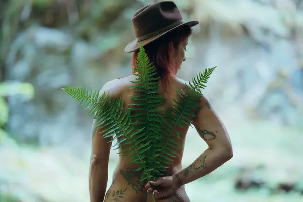 Sırtında yeşil eğrelti otu yaprakları ve kahverengi şapkalı kızıl saçlı kadın. Doğada ve arkadan fotoğraflanmış. Sağlık ve doğa projeleri kavramı.