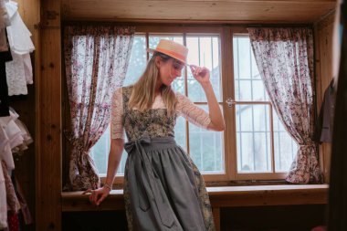 Şapka takan sarışın bir kadın ve Bavyera Dirndl elbisesi geleneksel bir kostüm dükkanının vitrininde duruyor..