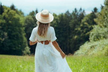 Sarışın bir kadının hasır şapka giydiği ve beyaz bir yaz elbisesi giydiği arka manzara. Doğada güzel bir kadın. Yaz havası.