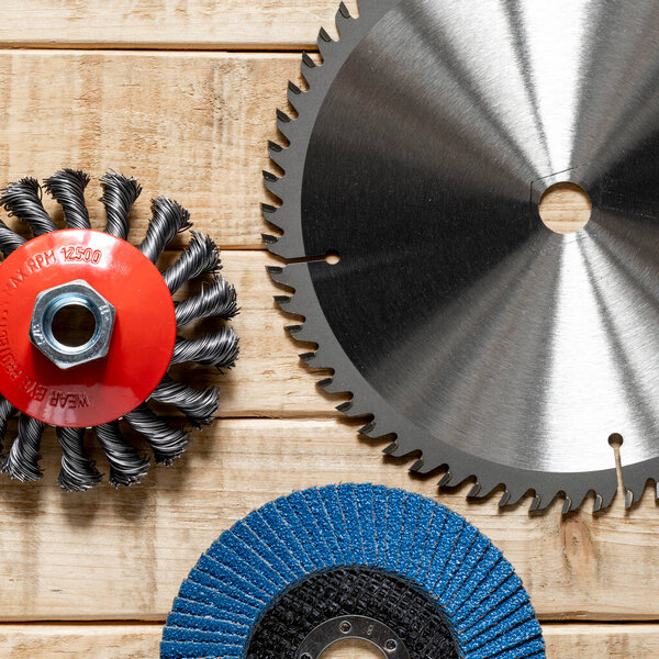 Three circular saw blades lay on a wooden surface. One is a wire brush, another is a toothed saw blade, and the last is a sanding disc.