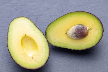 İki avokado yarım, birinin tohumu alınmış, diğerinin tohumu hala yerinde, gri bir arka planda yatıyor..