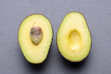 İki avokado yarım, birinin tohumu alınmış, diğerinin tohumu hala yerinde, gri bir arka planda yatıyor..