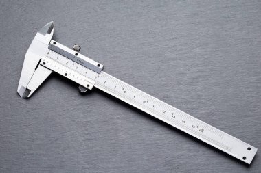 Üzerinde milimetre ve inç olarak siyah bir topuz ve işaretler olan gümüş kalibre gri bir levha yüzeyine yerleştirilmiştir. Caliper çene kapalı dik duruyor.