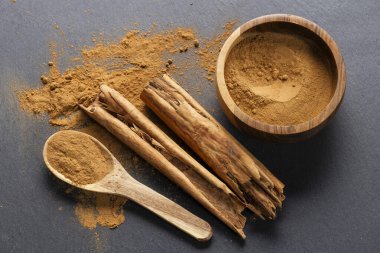 İki tarçın çubuğu gri bir tahta yüzeyde, etrafa saçılmış tarçın tozu ve tozla dolu tahta bir kaşık..