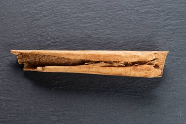 Koyu gri bir yüzeye dağılmış tarçın çubuklarının yakın plan görüntüsü. Tarçın parçaları açık kahverengidir, pürüzlü dokuları vardır ve bazıları kıvrımlıdır..