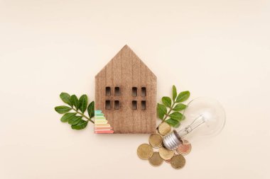 Yeşil yapraklı ahşap ev modeli, arkaplanda ampul ve madeni paralar evlerdeki enerji verimliliğini ve sürdürülebilirliği sembolize ediyor.