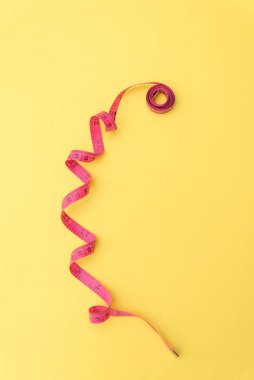 Sarı zemin üzerinde pembe mezura kıvrımlı. Dikiş, terzi ve vücut ölçüleriyle ilgili canlı ve anlamlı bir görüntü yaratıyor.