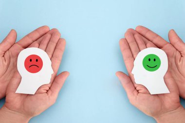 Mavi arka planda pozitif ve negatif emojili insan kafalarının kağıt kesiklerini tutan eller, pozitif ve negatif zihin veya müşteri geri bildirimi