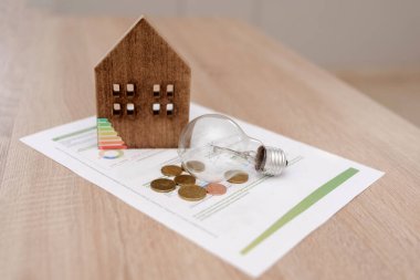 Wooden house model, light bulb and coins on energy efficiency document on the table clipart